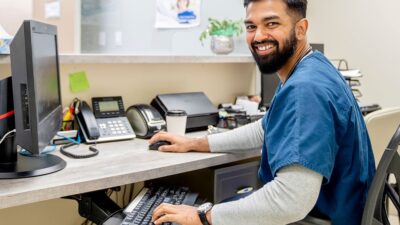 Smiling man wearing medical scrubs, working at his computer in a clinic.