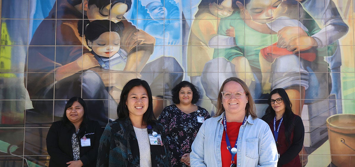 The team at Salud Para La Gente stands in front of a tile mural of motherhood.