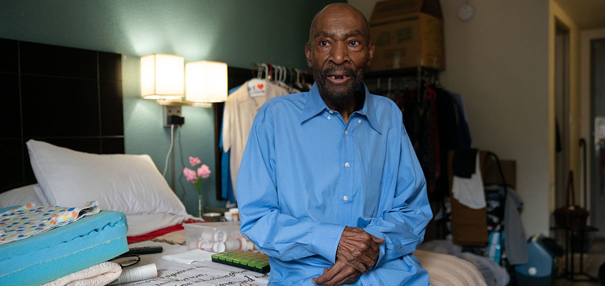 Elderly man sitting on hotel bed surrounded by his belongings.